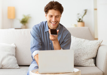 A man is sitting on a couch, smiling while looking at a smartphone in his hand. He appears to be using a voice assistant to complete a task while working on a laptop.