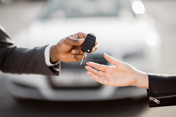 A man in a suit extends his hand to a woman in a leather jacket, holding a car key. The woman reaches her hand out to receive the key, cropped