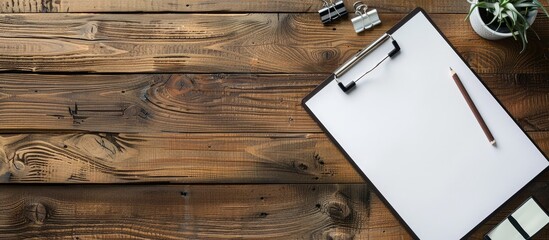 Fototapeta premium Aerial view of a wooden desk with a white clipboard holding blank paper, along with a pen, pencil, and clips. Ample space available for text or images.
