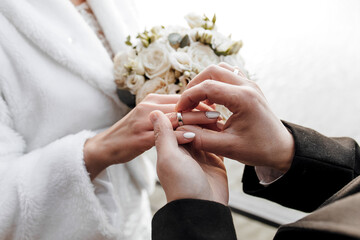 A bride and groom are getting married and exchanging rings. The bride is wearing a white coat and the groom is wearing a black suit. The bride is holding a bouquet of flowers