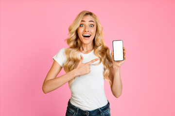 This image shows a young woman with long, blonde hair wearing a white t-shirt and blue jeans standing in front of a pink background. She is holding a smartphone with a blank screen