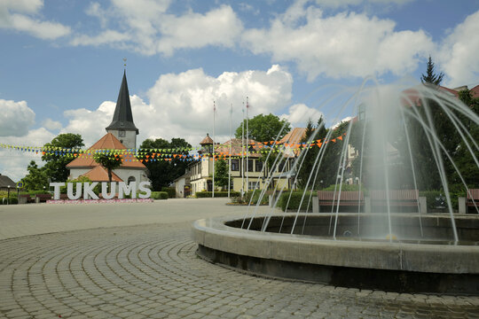 Main square in the centre of the town TUKUMS with its name on plinths. Latvia flags and bunting can also be seen behind a long exposure of the town fountain. Tukums, Latvia 06 04 2024 