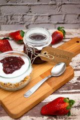 Glass jar full of muesli and yogurt with homemade strawberries jam, delicious snack with cheesecake and strawberries jam on wooden cutting board, detail vintage cardboard tag, vertical image social