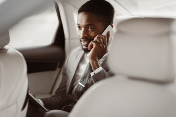 African American man manager in a suit sits in the backseat of a car and talks on his phone. He looks intently at the phone and appears to be engaged in a conversation.