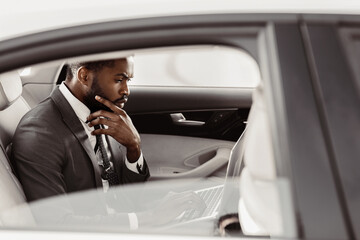 Black businessman, dressed in a suit and tie, sits in the backseat of a car, working on a laptop. He has a thoughtful expression on his face as he focuses on his work