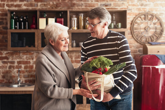 A senior couple stands in their kitchen, unpacking groceries from a reusable tote bag. The woman smiles warmly at her husband as he holds the bag, which contains fresh produce - Powered by Adobe