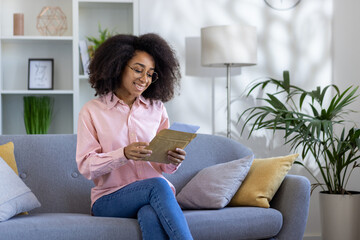 Smiling woman reading letter while sitting on cozy sofa in bright, modern living room. Happy moment of receiving good news or heartfelt message in comfortable home environment.