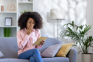 Woman wearing glasses reading shocking news in letter sitting on sofa in cozy living room. Reaction...
