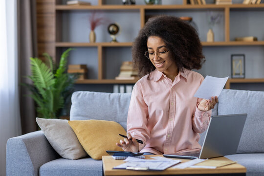Smiling woman managing finances at home using laptop, calculator, and documents in cozy living room. Concept of home budgeting, financial planning, and remote work