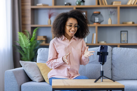 Woman in glasses sitting on sofa having video call on smartphone. She is smiling and gesturing. Used for online communication, remote work, social media, or video chat