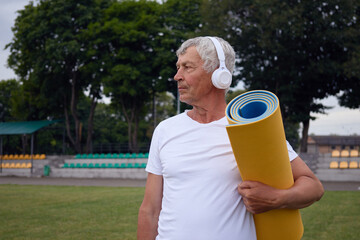 Serious Caucasian mature man holding yoga mat and looking away having fitness exercise at the stadium listening music or online meditation while having workout