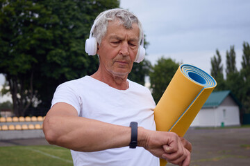 Serious senior man wearing white T-shirt holding yoga mat using smart watch looking at heart rate monitor old man using technology for health and sport activity
