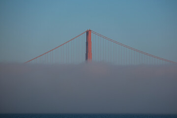 Obraz premium World famous red steel suspension bridge in San Francisco port entrance with Golden Gate State park nature lands hidden in cloud mist fog bank with clear sky pilars