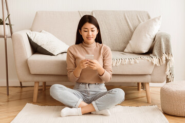 A young woman in a beige sweater and blue jeans sits on the floor in front of a couch, using her smartphone.