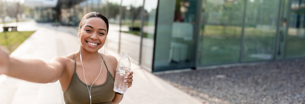 African American woman taking selfie with cellphone, recording video for her blog, holding water bottle on street. Black lady influencer communicating to followers, copy space