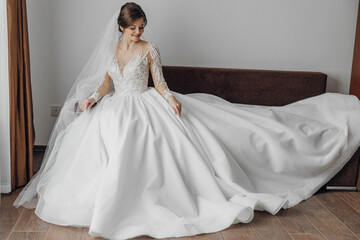 Fototapeta premium A woman in a white wedding dress is sitting on a couch. The dress is long and flowing, and the woman is wearing a veil. Concept of elegance and celebration
