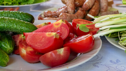 Ripe bright red sliced ​​tomatoes, cucumbers and greens close-up against background of delicious fried sausages on table
