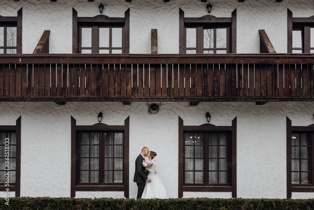 Wall mural A bride and groom are kissing in front of a building with brown shutters. The building is white and has a rustic feel