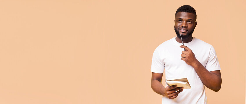 A young black man with a beard is thoughtfully holding a notebook and pen in front of a beige background. He is looking off to the side, and his facial expression suggests that he is deep in thought.