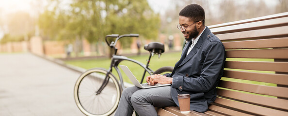 Obraz premium An African American man works on his laptop while sitting on a park bench, his bicycle resting nearby. The setting is peaceful and productive, combining work with the outdoors.