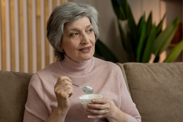 A video of an elderly woman eating yogurt while sitting on a couch in her living room. The woman is dressed in a pink sweater and the room is lit by warm lamps.