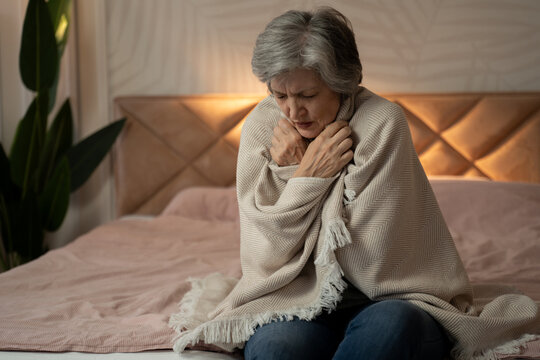 A woman in her late 60s or early 70s is sitting on a bed, wrapped in a blanket, and looking unwell. She appears to be cold and may be feeling sick