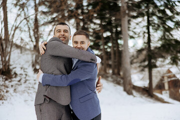 Two men hug each other in the snow. One of them is wearing a blue jacket. Scene is warm and friendly