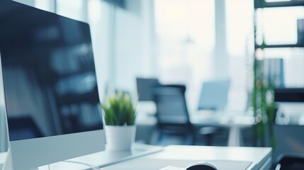 A white desktop computer sits on a desk in an office setting, with a blurred background of other desks and windows. The computers screen is turned off, and a small potted plant sits beside it