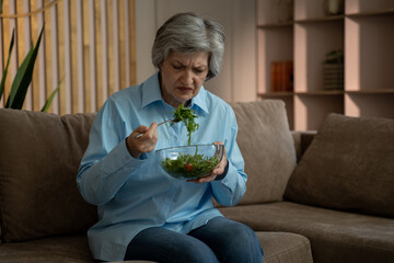 An elderly woman is sitting on the couch in the living room. She's eating a salad, but you can tell...