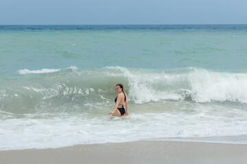 A woman in a black swimsuit stands in the shallows of a beach, smiling as a wave rolls in around her