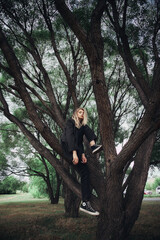 A woman sits on a tree branch in a forest. She is wearing black pants and a black jacket. Her blonde hair is flowing in the wind. The trees are tall and the day is cloudy