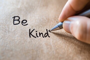 Close-up of a hand using a pen to write "Be Kind" on textured paper, emphasizing a message of kindness and positivity.