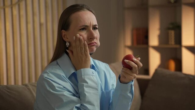Woman experiencing a toothache while eating an apple in her living room. She appears to be in pain and grimaces as she chews