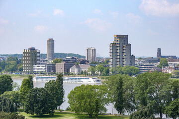 Rhine river with a tourist boat seen from high perspective, green leafed trees of Rheinpark public park in foreground, cityscape with buildings in background, cloudy spring day in Cologne, Germany