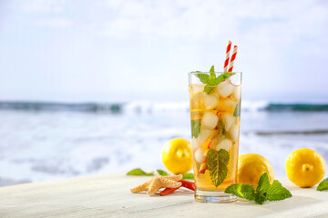 Image of copy space for drinks and food and products. Glass of mineral water with lemon slices, ice and straws on white table with blurred seascape and rocky mountains background.