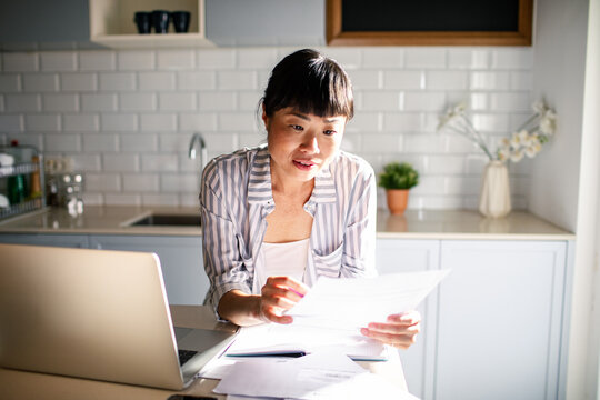Asian woman reading bills in the kitchen with laptop at home