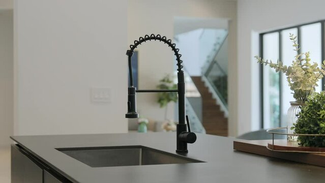 Modern kitchen interior. Focus on sleek, black faucet over sink that built into granite countertop. Background of blurred view of bright, airy room with large windows