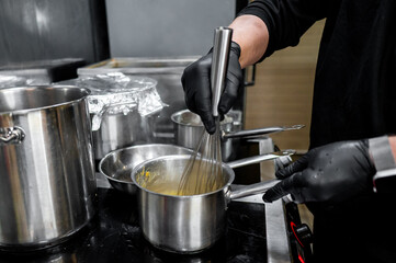 A close-up of a hand wearing a black glove, whisking in a silver pot on a stove, depicting cooking or food preparation