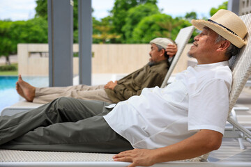 Two senior asian man are sitting at the beach bed by swimming pool in the tropical resort for retirement vacation and luxury leisure recreation travel concept