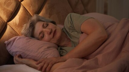 At night, an elderly woman can be seen sleeping soundly in bed. She is covered with a blanket, and her arms are wrapped around a pillow. The bed is made with pink sheets and a pink blanket