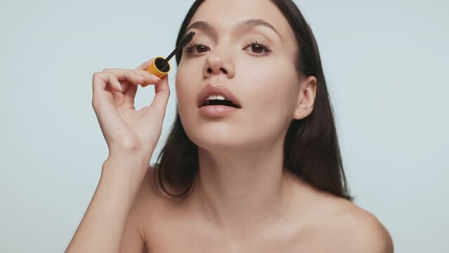 Attractive young brunette woman carefully applying mascara, looking into the camera against a light blue studio background. Beauty and makeup concept.