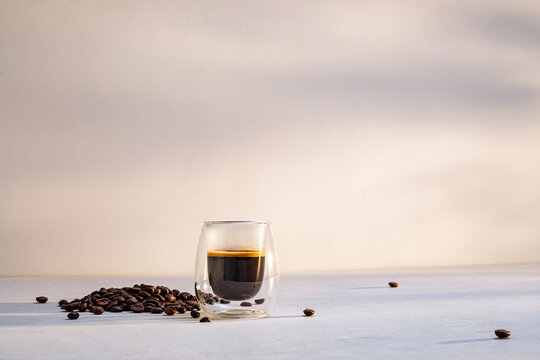 A glass of dark espresso sits on a white tabletop, surrounded by scattered coffee beans in the morning.