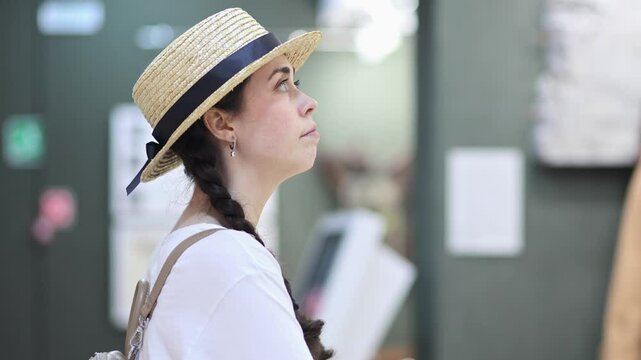 Exhibition in gallery. Side view of young Caucasian woman wearing straw hat contemplating exposition. Cultural education excursion for students and Museum's Day