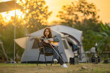 Confident young woman with interested facial expression playing acoustic guitar and watching for stunning mountain scenery from her tent set up on green lawn and beautiful sunset above mountains.