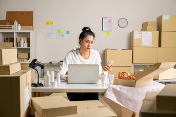 Asian e-commerce seller wating for order confirmations via smartphone and laptop in a warehouse...