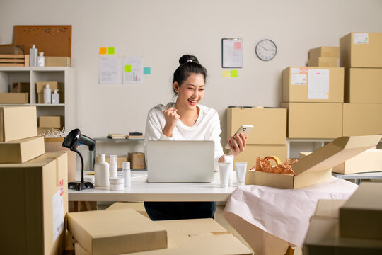Asian e-commerce seller receives order confirmations via smartphone and laptop in a warehouse setting for an online beauty shop.
