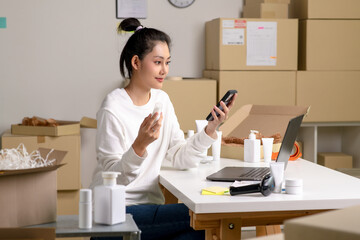 Asian e-commerce seller receives order confirmations via smartphone and laptop in a warehouse setting for an online beauty shop.
