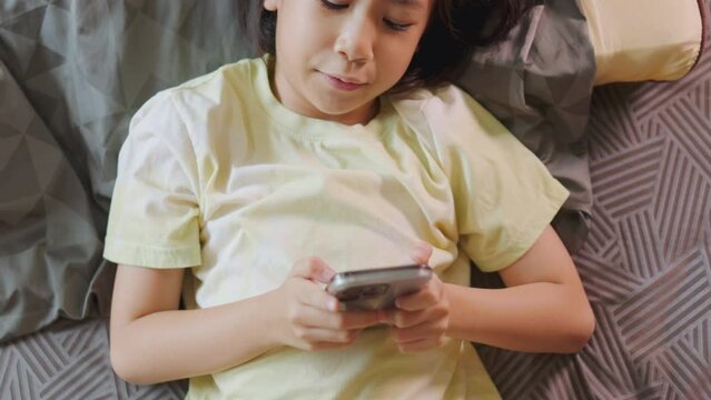 Top view of adorable Asian kid girl, lied in bed, enjoys her smartphone in a sunlit room. Her expression hints at digital fun, highlighting the allure of home tech, playing game and e-learning.
