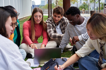 Multiracial group of students gathered outdoors in the university campus, sitting on the grass studying together and doing homework using a laptop. 
