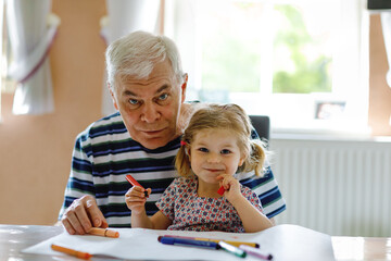Cute little baby toddler girl and handsome senior grandfather painting with colorful pencils at...